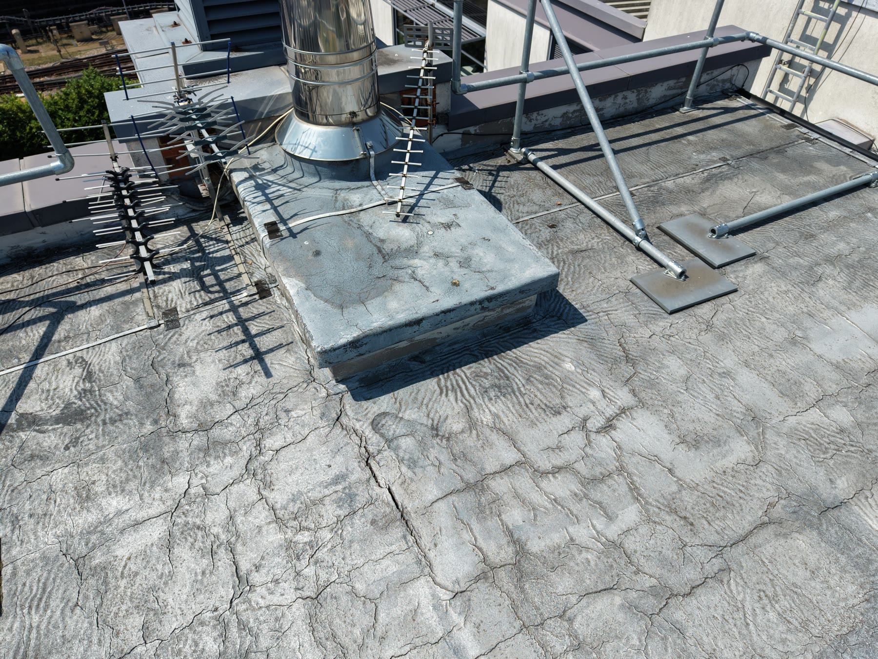 Close-range drone roof inspection showing cracked flat roof membrane around a flue and antenna base